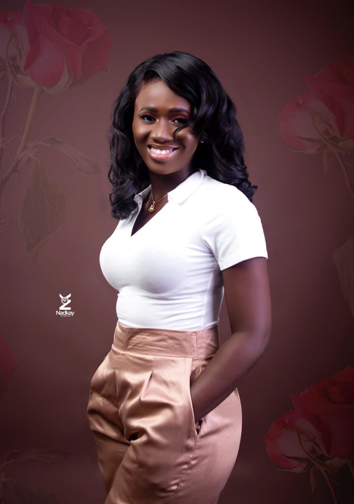 A smiling young woman wearing a white top and beige pants, standing confidently with her hands in her pockets against a backdrop of roses. Healing after heartbreak with God