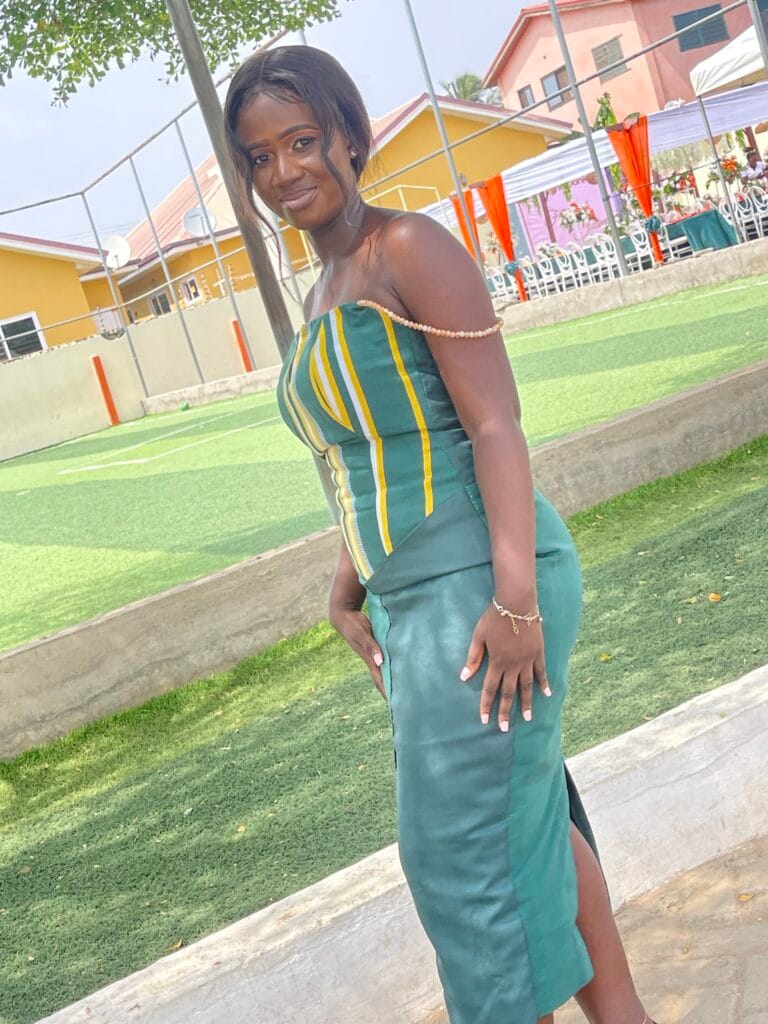 A young woman standing outdoors, smiling while wearing a stylish green and striped dress, with vibrant buildings and a sports court in the background. Daisy is Healing after heartbreak with God.