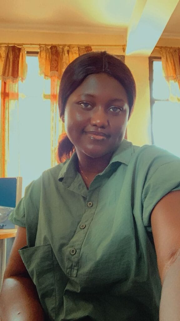 A woman smiling while wearing a green shirt, seated in a room with natural light coming through the windows and curtains in the background.