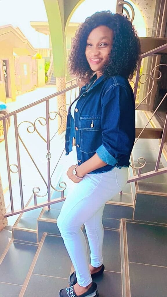 A woman confidently standing on a balcony, wearing a denim jacket and white pants, with a smile that exudes grace and positivity. She put God to the test.