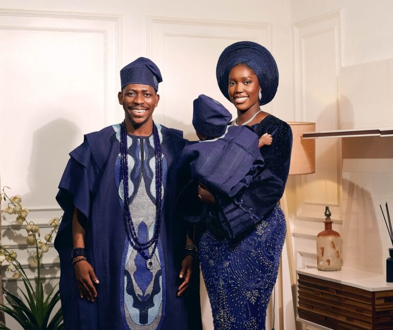 Moses Bliss and his wife, Marie Bliss, pose joyfully indoors in matching navy blue traditional attire, holding their newborn son dressed in similar clothing.