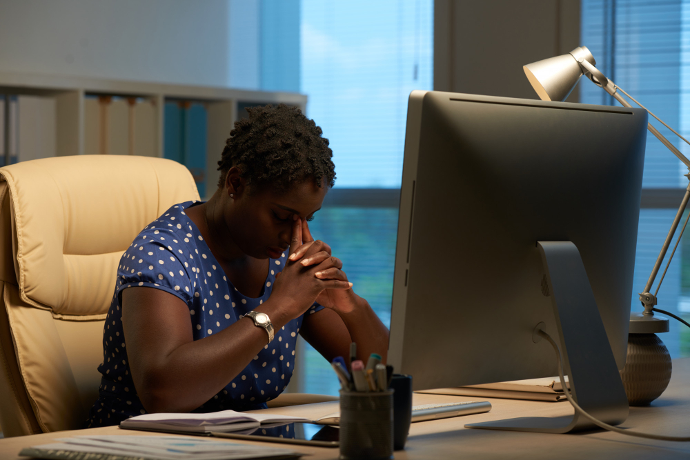 Afro-american woman sitting in front of a computer in an office and leaning her head on clasped hands. Building her spiritual life while busy.