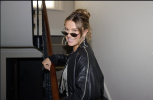 Millie Bobby Brown smiling confidently on a staircase, leather jacket and sunglasses, playful energy