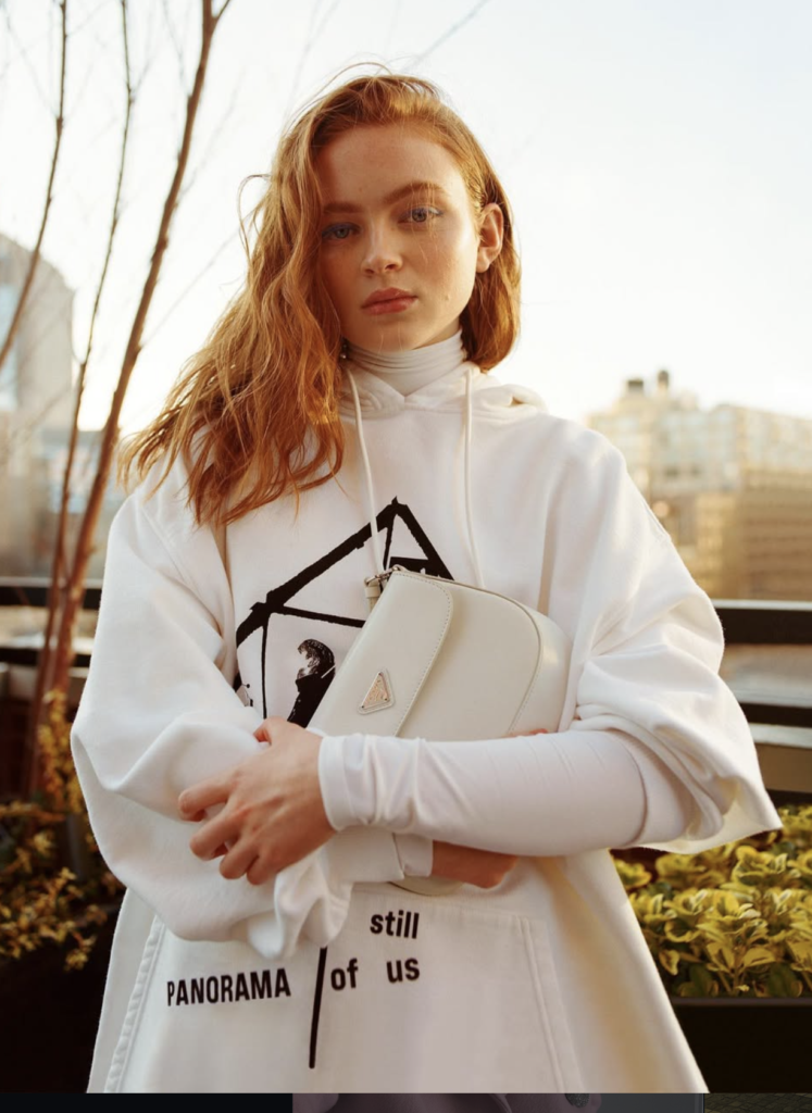 Young red-haired woman in white hoodie outdoors holding handbag, calm serious expression