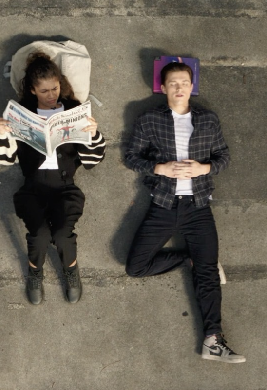 Zendaya reading a newspaper while Tom Holland lies beside her in a relaxed Spider-Man scene.