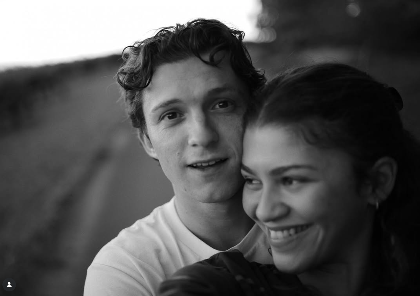 Zendaya smiling while Tom Holland stands close behind her in a candid black and white photo