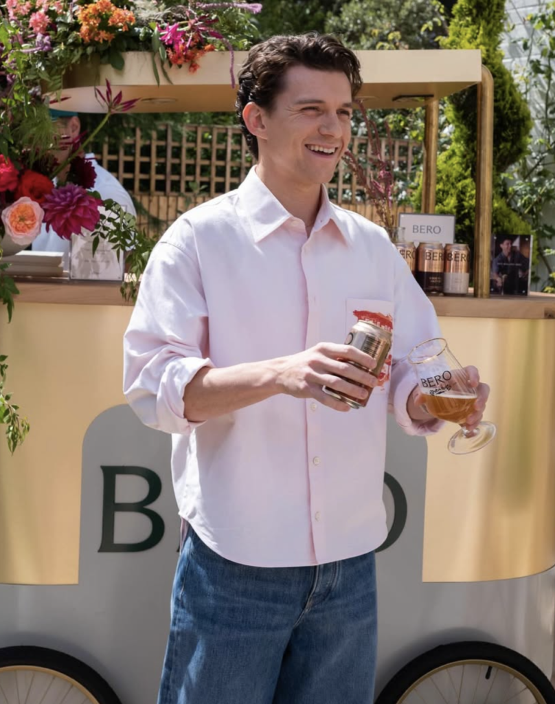 Tom Holland smiling outdoors while holding a drink at a branded event, dressed in a light pink shirt and jeans