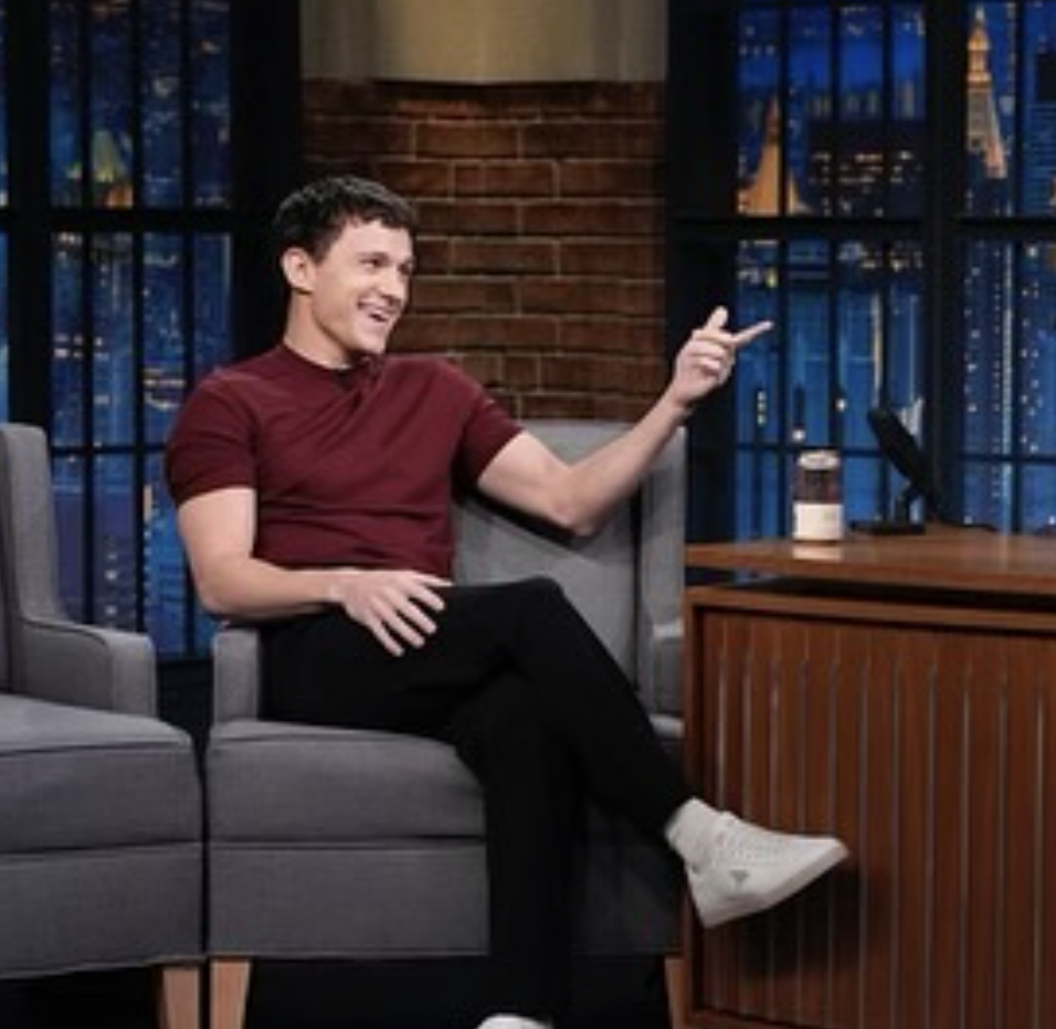 Tom Holland sitting on a talk show set smiling and pointing, wearing a maroon shirt and white sneakers
