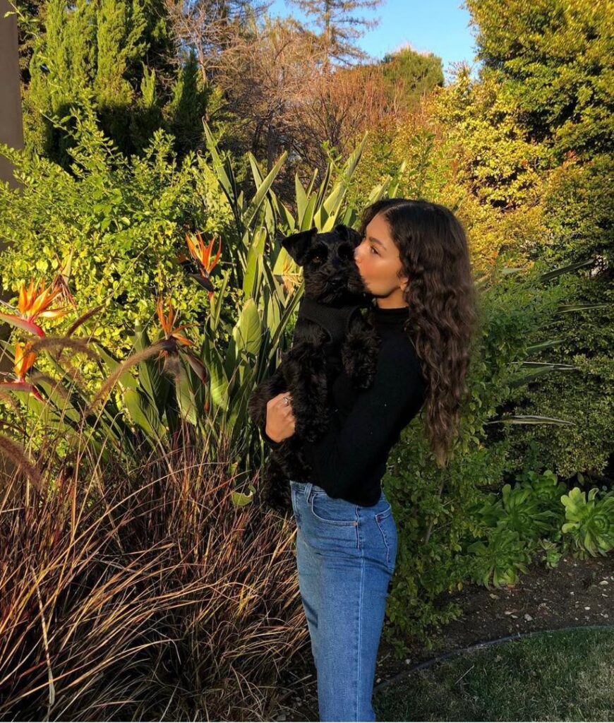 Zendaya holding a black dog in a peaceful garden surrounded by greenery and flowers.