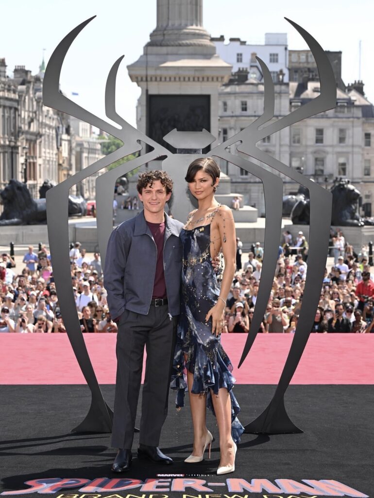 Tom Holland and Zendaya pose together in front of the giant Spider-Man logo during the Spider-Man: Brand New Day press tour.