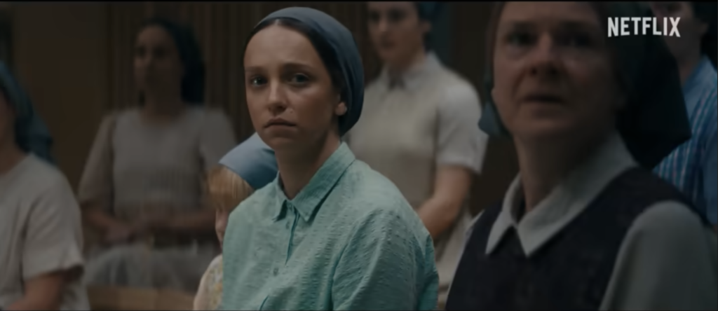 Rosie sits quietly among members of a strict religious community in The Unchosen, appearing thoughtful and concerned.
