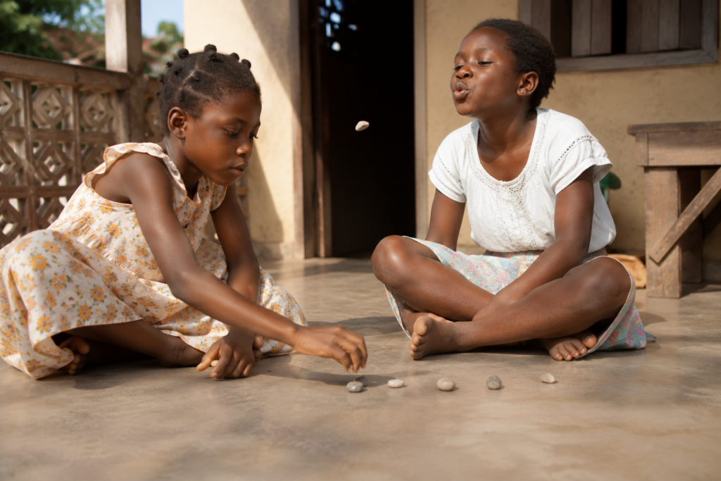 Two young Ghanaian sisters play the traditional En-Ones game with seven small stones on a cement veranda while one playfully blows air to distract the other.