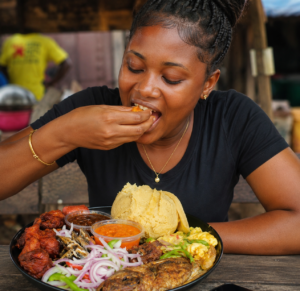 A smiling young Ghanaian woman enjoying Ga kenkey with fried fish, pepper sauce, shito, onions, and traditional side dishes.