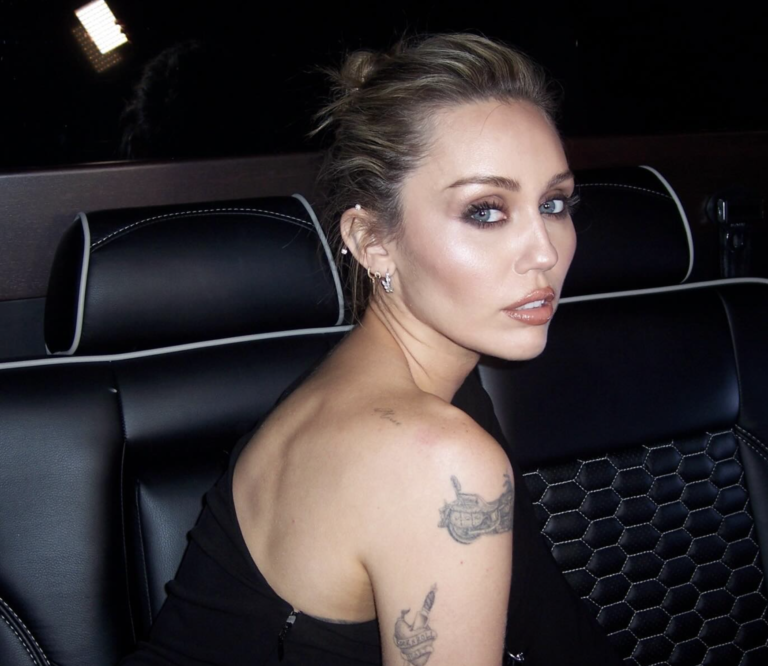 Miley Cyrus looking over her shoulder while seated in a car wearing a black dress.