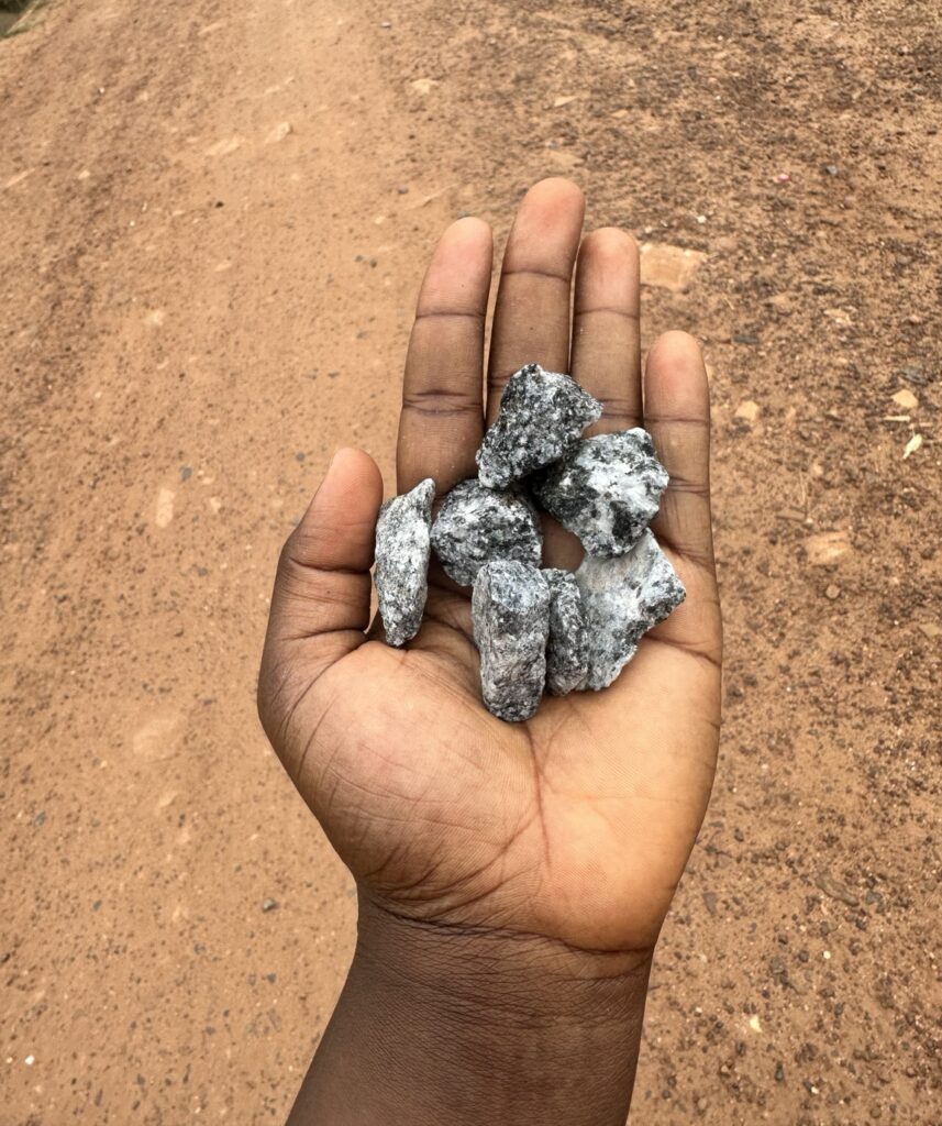 A hand holding seven small stones on a dusty road in Ghana, reminding the author of the traditional Ghanaian childhood game En-Ones.