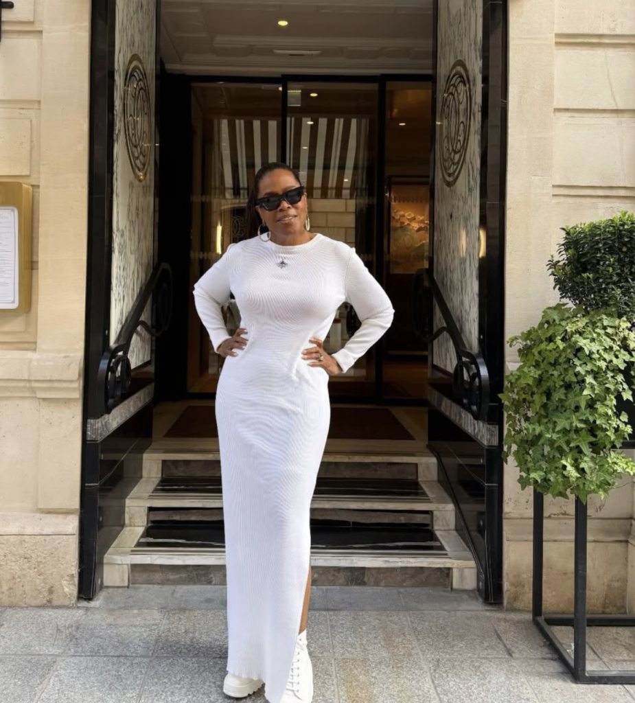 Oprah Winfrey standing outside a building in a white dress and sunglasses